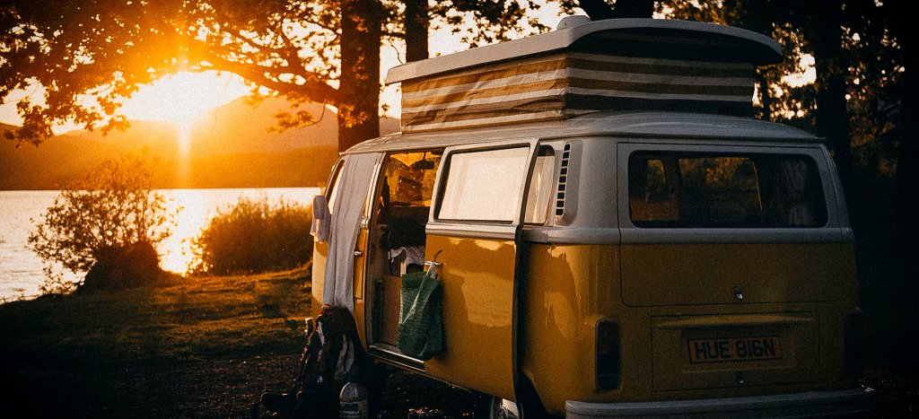 En klassisk gul campervan med uppfällt tak parkerad vid en sjö i solnedgången, symboliserar friheten med vanlife.